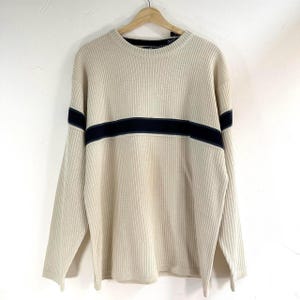 May include: A cream-colored sweater with a black stripe across the chest. The sweater is made of a soft, ribbed knit fabric.
