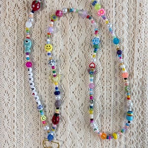 May include: A long lanyard made of multicoloured beads in various shapes and sizes, including smiley faces, flowers, cherries, a ladybug, and a fish. The beads spell out NIKKARN with a heart symbol. A gold-toned metal lobster clasp is at the end.