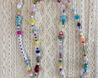 Puede incluir: Un cordón largo hecho de cuentas multicolores de varias formas y tamaños, incluyendo caras sonrientes, flores, cerezas, una mariquita y un pez. Las cuentas deletrean NIKKARN con un símbolo de corazón. Un mosquetón de metal dorado está en el extremo.