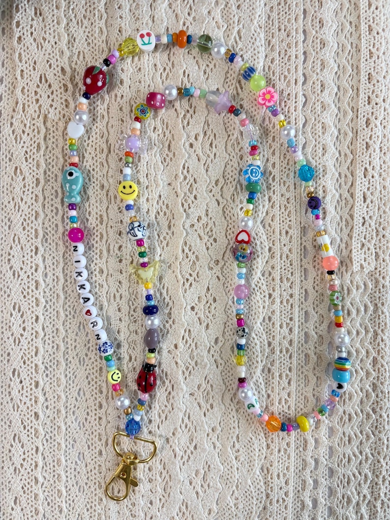 May include: A long lanyard made of multicoloured beads in various shapes and sizes, including smiley faces, flowers, cherries, a ladybug, and a fish. The beads spell out NIKKARN with a heart symbol. A gold-toned metal lobster clasp is at the end.