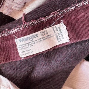 May include: Close-up of a pair of burgundy Wrangler jeans. The tag reads "Wrangler" with size 33x32. The tag also includes washing instructions and the material composition, 100% cotton. The jeans are made in the USA.