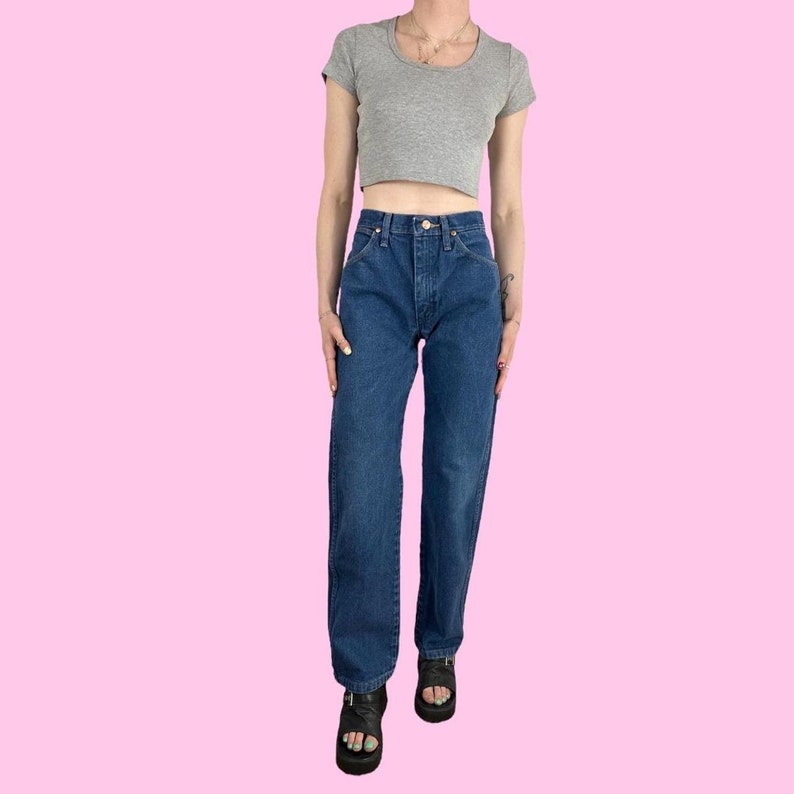 Puede incluir: Un par de jeans azules de tiro alto y pierna recta con una camiseta gris claro recortada. La modelo lleva sandalias negras.