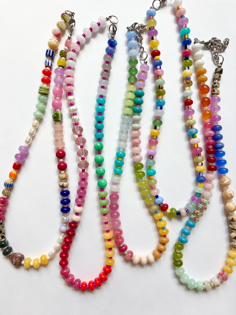 May include: A collection of colorful beaded necklaces. Each necklace features a unique combination of beads in various shapes, sizes, and colors, including pink, blue, green, yellow, and red. The necklaces have silver clasps and are displayed against a white background.
