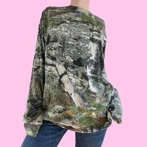 May include: A long-sleeved shirt with a camouflage pattern featuring a woodland scene. The shirt is green, brown, and white.