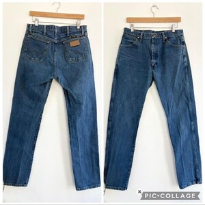 May include: Two pairs of blue denim jeans hanging on wooden hangers. The jeans have a classic five-pocket design, with a brown leather patch on the back. The jeans are a medium wash and appear to be vintage.