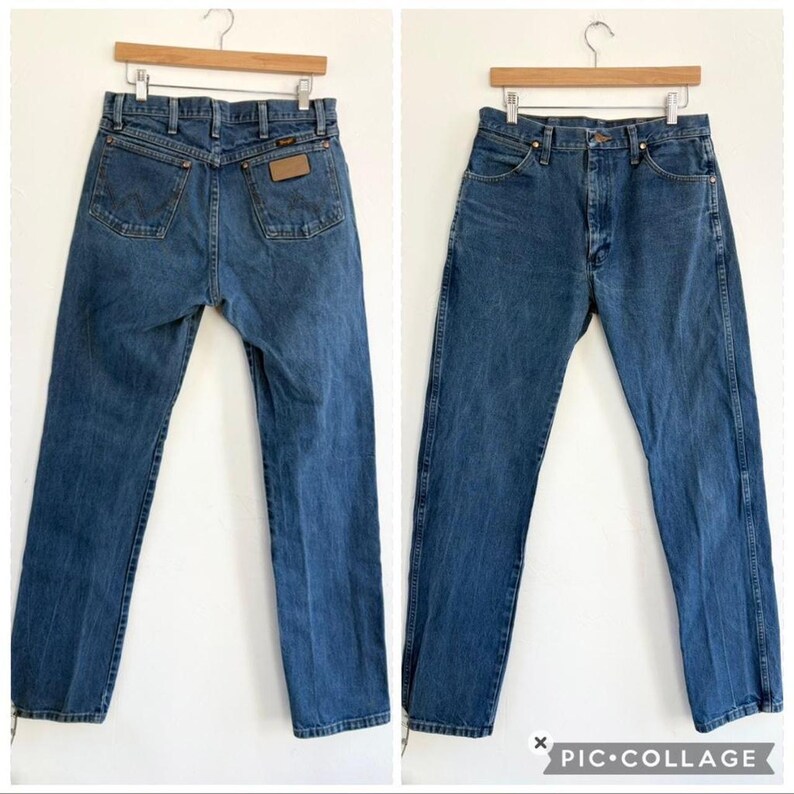 May include: Two pairs of blue denim jeans hanging on wooden hangers. The jeans have a classic five-pocket design, with a brown leather patch on the back. The jeans are a medium wash and appear to be vintage.