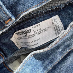 May include: Close-up of blue denim jeans with a white tag. The tag reads "Wrangler" and includes size information: 32 x 38. The tag also provides washing instructions and fabric details. The jeans are made of US fabric.