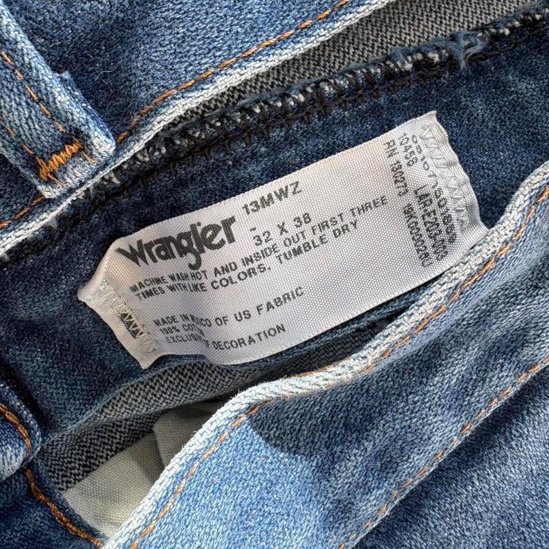 May include: Close-up of blue denim jeans with a white tag. The tag reads "Wrangler" and includes size information: 32 x 38. The tag also provides washing instructions and fabric details. The jeans are made of US fabric.