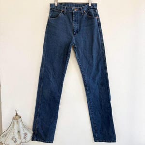 May include: Dark blue denim jeans hanging on a metal hanger. The jeans have a classic five-pocket design with a button and zipper closure. The denim appears to be a medium weight, suitable for everyday wear. The jeans are straight-legged.