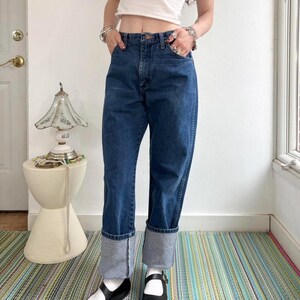 May include: Dark wash denim jeans with a cuffed hem. The jeans have a classic five-pocket design and a button and zipper closure. The cuffs are a lighter shade of blue. The person is wearing black shoes.