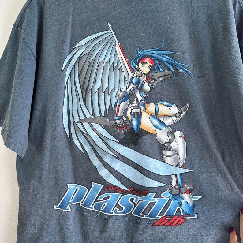 May include: A blue t-shirt with a graphic print of a female anime character with wings, holding a gun. The text "Plastik" is printed below the character in Japanese and English.