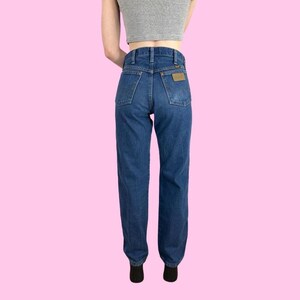 Puede incluir: Un par de pantalones vaqueros de mezclilla azul oscuro con un parche de cuero marr&oacute;n en el bolsillo trasero. Los pantalones tienen cintura alta y pierna recta.
