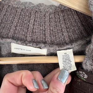 May include: A brown sweater with a tag that reads "sleeping on snow" and a smaller tag that lists the fabric composition and size as "XS". The fabric composition is 61% wool, 15% nylon, 8% acrylic, 7% angora rabbit hair, 7% polyester, and 2% spandex. The sweater is made in China.