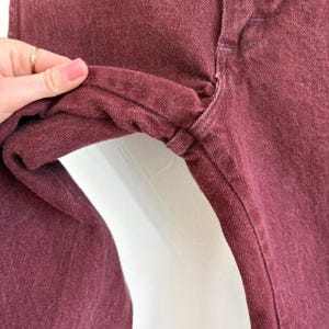 May include: Close-up of burgundy-colored denim jeans. The fabric is a solid color with visible stitching and seams. The jeans are folded, showing the texture and construction of the garment.