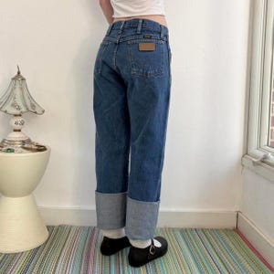 May include: Dark wash denim jeans with a brown leather patch on the back pocket. The jeans have a straight leg with a folded cuff at the hem. The person is wearing black Mary Jane shoes.
