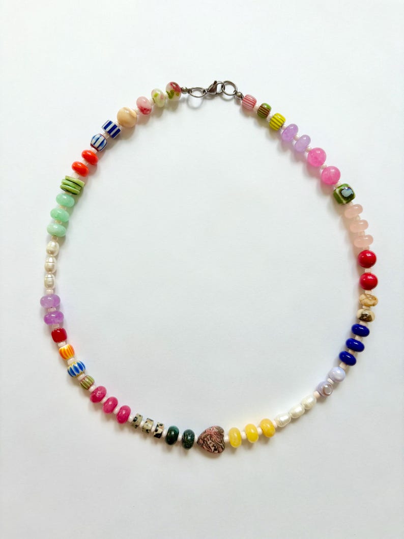 May include: A colorful beaded necklace featuring a variety of shapes and sizes. The necklace includes beads in shades of pink, red, yellow, blue, green, and white, with a heart-shaped bead and a silver clasp.