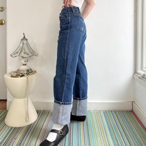 May include: Blue denim jeans with a light blue cuff. The jeans are worn with black Mary Jane shoes with a small buckle. The person is standing on a striped rug.