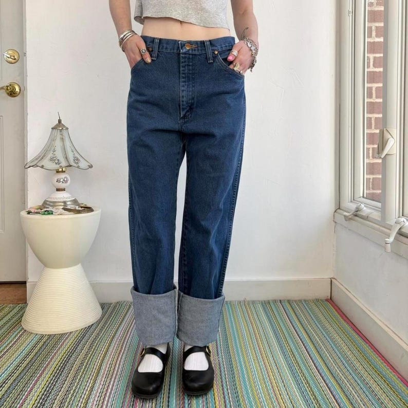 May include: Dark wash denim jeans with cuffed hems. The jeans have a classic five-pocket design and a straight leg fit. The model is wearing black Mary Jane style shoes. The jeans are a medium-dark blue color.