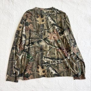 K&ouml;nnte beinhalten: Lang&auml;rmeliges Camouflage-Shirt in Braun-, Gr&uuml;n- und Beiget&ouml;nen. Das Shirt hat einen Rundhalsausschnitt und ein Waldmuster, ideal f&uuml;r Outdoor-Aktivit&auml;ten oder die Jagd.