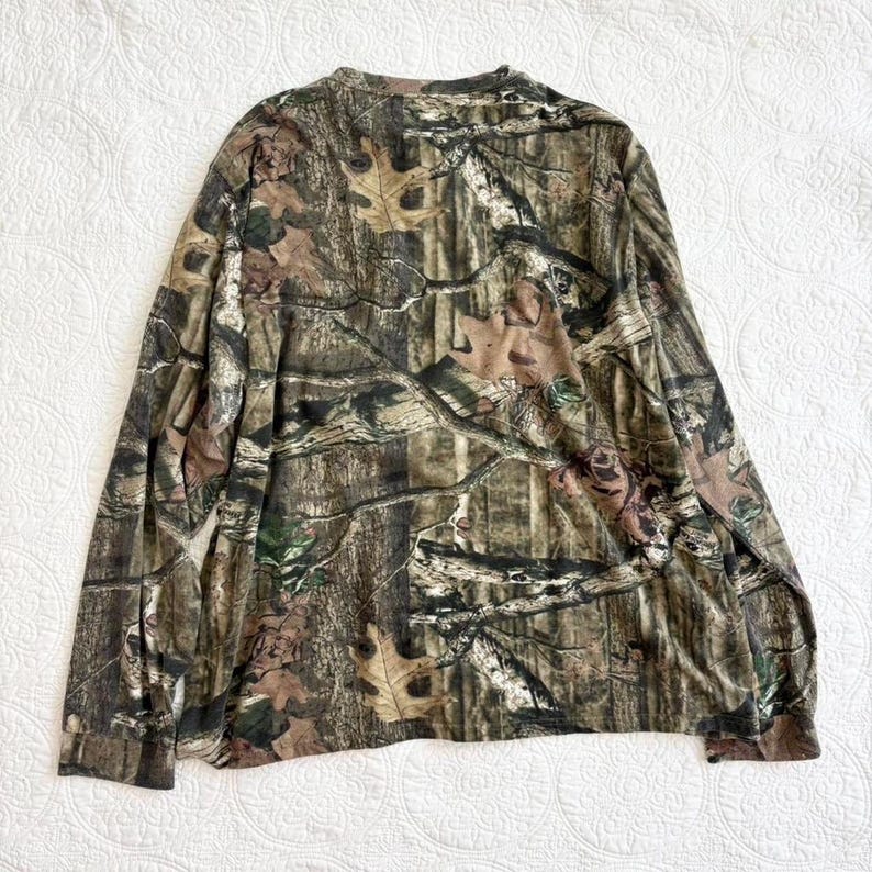 K&ouml;nnte beinhalten: Lang&auml;rmeliges Camouflage-Shirt in Braun-, Gr&uuml;n- und Beiget&ouml;nen. Das Shirt hat einen Rundhalsausschnitt und ein Waldmuster, ideal f&uuml;r Outdoor-Aktivit&auml;ten oder die Jagd.