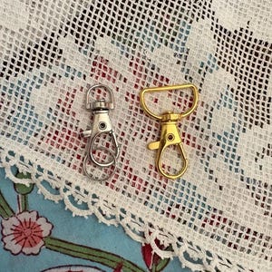 May include: Two metal swivel lobster claw clasps. One clasp is silver-coloured with a circular attachment ring at the top, and the other is gold-coloured with a D-shaped attachment loop at the top. Both feature a spring-loaded gate mechanism.