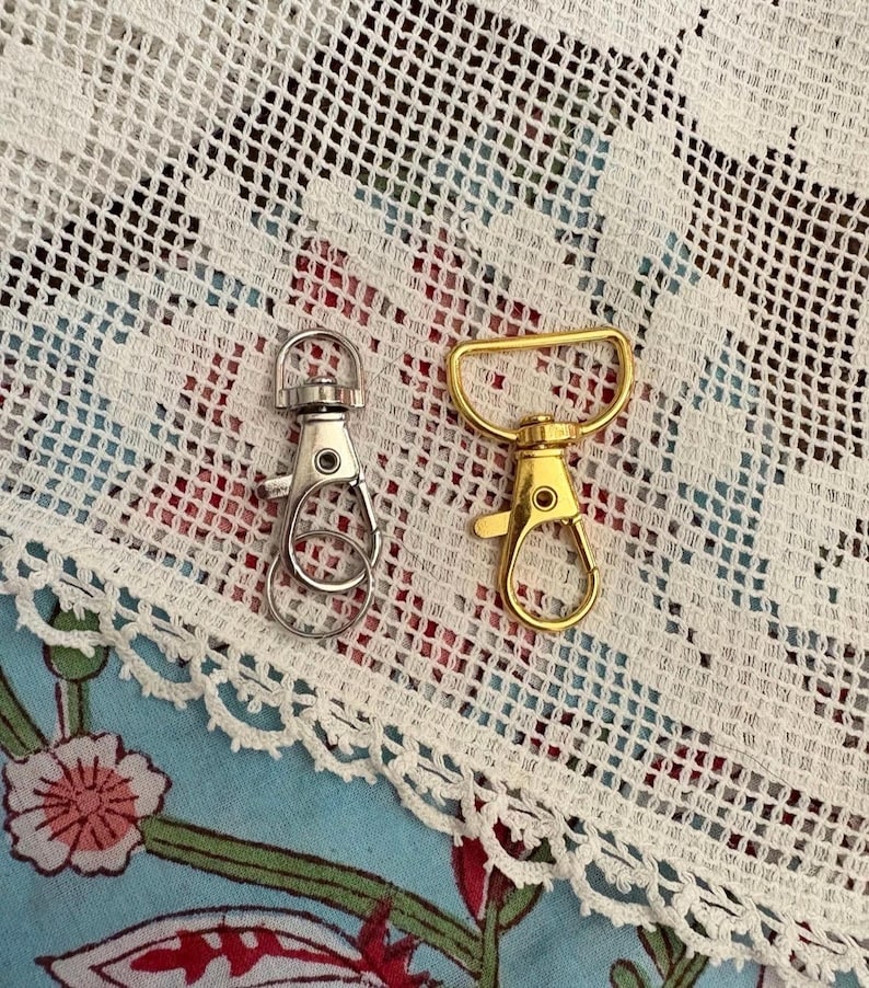 May include: Two metal swivel lobster claw clasps. One clasp is silver-coloured with a circular attachment ring at the top, and the other is gold-coloured with a D-shaped attachment loop at the top. Both feature a spring-loaded gate mechanism.