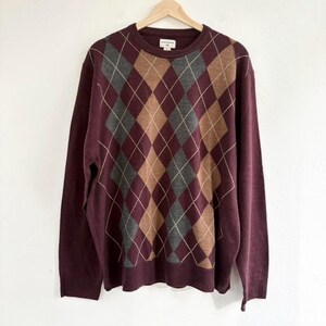 May include: A burgundy argyle sweater with a crew neck, long sleeves, and a classic diamond pattern in shades of burgundy, brown, gray, and white. The sweater is hanging on a wooden hanger.