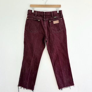 May include: Burgundy-colored denim jeans with a classic five-pocket design. The jeans have a straight leg cut and a frayed hem. The Wrangler brand label is visible on the back pocket. The jeans are hanging on a wooden hanger.