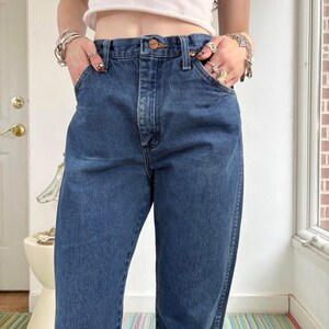 May include: Dark wash denim jeans with a classic five-pocket design. The jeans have a high waist and a relaxed fit. The model is wearing the jeans with their hands in the pockets. The jeans are a medium blue color.
