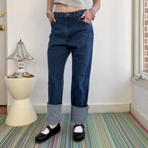 May include: Dark blue denim jeans with cuffed hems. The jeans have a classic five-pocket design and a straight leg fit. The cuffs are a lighter shade of blue. The person is wearing black Mary Jane shoes.