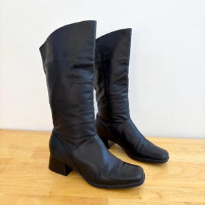 May include: A pair of black leather knee-high boots with a block heel. The boots have a square toe and a slouchy fit.