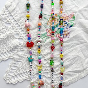 May include: Two beaded lanyards featuring a variety of colourful plastic and glass beads in shapes including animals, fruits, flowers, and smiley faces. One lanyard includes the text VET TECH and the other includes the name ALLIE, both with gold clasps.