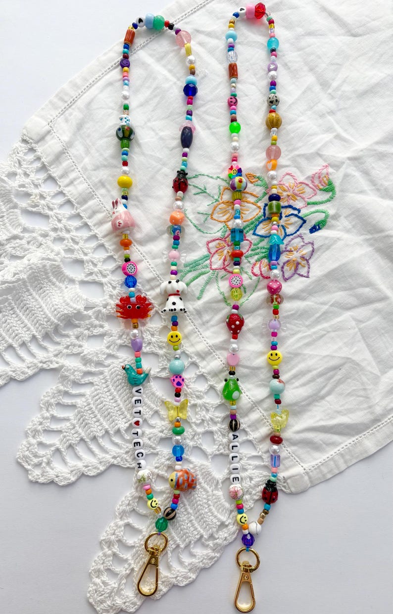 May include: Two beaded lanyards featuring a variety of colourful plastic and glass beads in shapes including animals, fruits, flowers, and smiley faces. One lanyard includes the text VET TECH and the other includes the name ALLIE, both with gold clasps.