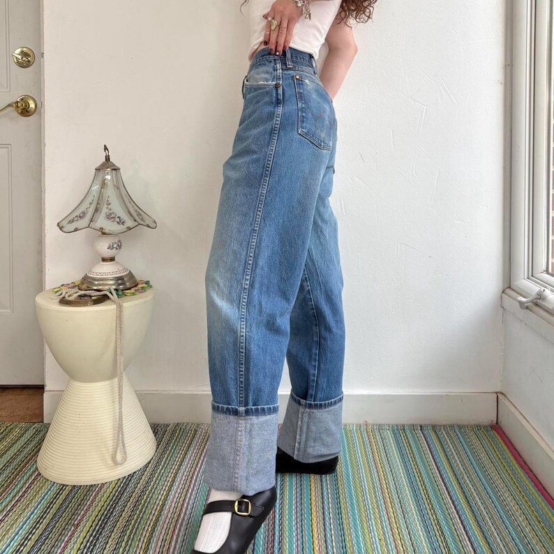 May include: Light blue denim jeans with a cuffed hem. The jeans have a classic five-pocket design and a relaxed fit. The model is wearing black shoes with a white sock. The jeans are a medium wash and appear to be vintage.