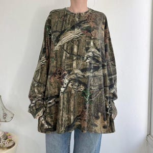 K&ouml;nnte beinhalten: Lang&auml;rmeliges Camouflage-Shirt in Braun-, Gr&uuml;n- und Schwarzt&ouml;nen. Das Shirt hat einen Knopfleisten-Ausschnitt und eine lockere Passform. Der Stoff hat ein strukturiertes Aussehen, und das Muster ahmt eine Waldumgebung nach.