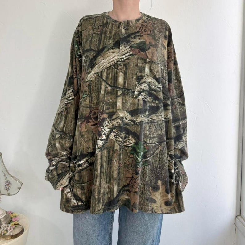 K&ouml;nnte beinhalten: Lang&auml;rmeliges Camouflage-Shirt in Braun-, Gr&uuml;n- und Schwarzt&ouml;nen. Das Shirt hat einen Knopfleisten-Ausschnitt und eine lockere Passform. Der Stoff hat ein strukturiertes Aussehen, und das Muster ahmt eine Waldumgebung nach.