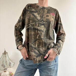 May include: Long-sleeved camouflage shirt in shades of brown, green, and tan. The shirt features a crew neck and a small pink logo on the upper chest. The fabric has a mottled pattern, typical of camouflage designs.