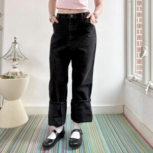 May include: Black denim jeans with turned-up cuffs. The jeans have a high waist and a straight leg. The person is wearing black Mary Jane shoes and white socks. The background includes a striped rug and a window.