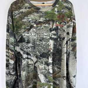May include: A long-sleeved shirt with a camouflage pattern featuring a green, brown, and white color scheme. The shirt has a crew neck and the Mossy Oak logo is visible on the left chest.