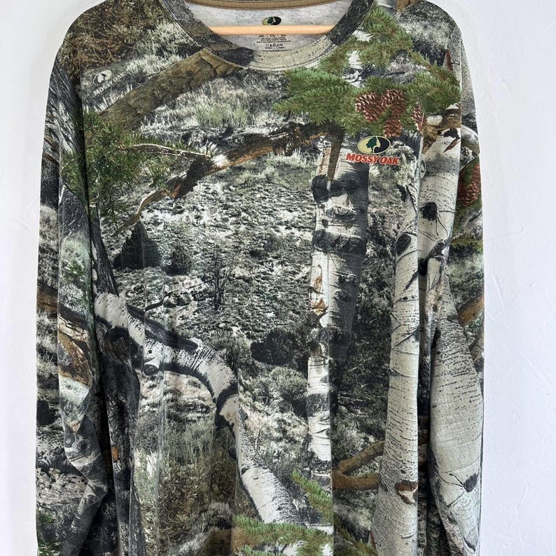May include: A long-sleeved shirt with a camouflage pattern featuring a green, brown, and white color scheme. The shirt has a crew neck and the Mossy Oak logo is visible on the left chest.
