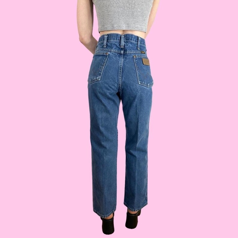 May include: Blue denim jeans with a high waist and a straight leg. The jeans have classic five-pocket styling and a brown leather patch on the back pocket. The jeans are worn with a gray crop top and black shoes. The background is a solid pink.