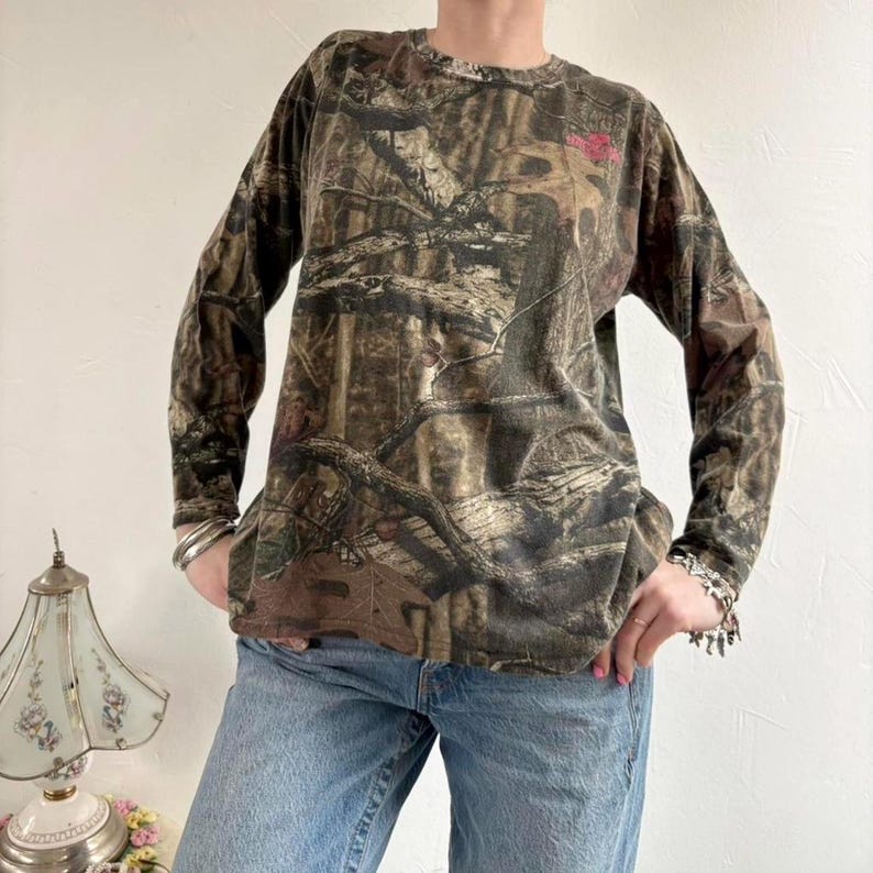 May include: Long-sleeved camouflage shirt in shades of brown, green, and tan. The shirt features a crew neck and a subtle pink emblem on the chest. The fabric has a dense, natural pattern, typical of hunting apparel.