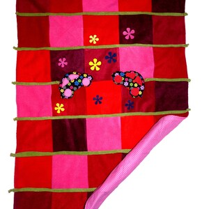 Könnte beinhalten: Ein Patchwork-Quilt mit roten, rosa und burgunderfarbenen Quadraten. Grüne Bänder akzentuieren die Nähte. Der Quilt hat Blumenapplikationen und eine rosa gepunktete Rückseite. Eine gemütliche, handgemachte Decke.