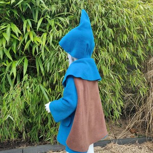 Könnte beinhalten: Ein Kinderkostüm mit einem blauen Kapuzenmantel, einer passenden spitzen Mütze und einem braunen Umhang. Das Outfit aus weichem Stoff ist als Zwerg- oder Zauberer-Outfit gestaltet. Der Hintergrund ist grüner Bambus.