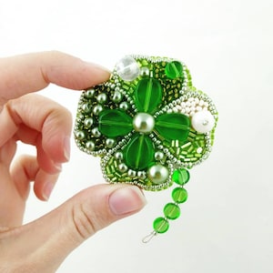 May include: A green four-leaf clover brooch made of beads and sequins. The clover is surrounded by white beads and has a green chain attached.