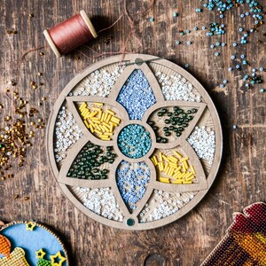 May include: A wooden bead organiser with a flower design. The organiser has seven compartments filled with different coloured beads. The beads are in shades of white, blue, green, and yellow.
