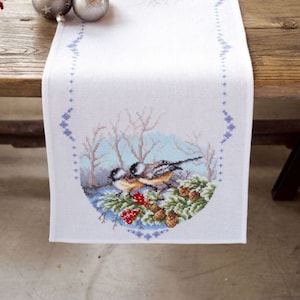 May include: White table runner with a cross-stitch design of two birds in a winter scene. The runner is on a wooden table with silver ornaments and a red berry branch.