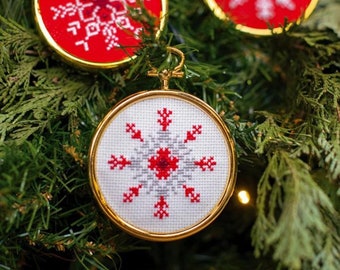 May include: Three red and white snowflake ornaments with gold frames. The ornaments are hanging on a green Christmas tree.