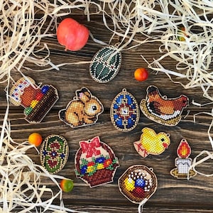 Può includere: Una collezione di 10 ornamenti a punto croce a tema pasquale. Gli ornamenti sono in legno e presentano una varietà di disegni, tra cui un coniglio, un pulcino, un cesto di uova e una candela. Gli ornamenti sono disposti su una superficie di legno con paglia.