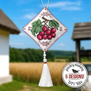 Könnte beinhalten: Ein rautenförmiges Kreuzstich-Ornament mit einer Traube aus roten Weintrauben und grünen Blättern auf weißem Grund, umrandet mit roten Stichen. Unten hängt eine weiße Quaste und ein rundes Abzeichen trägt die Aufschrift Jewelry making 5 DESIGNS! DIY Kits.
